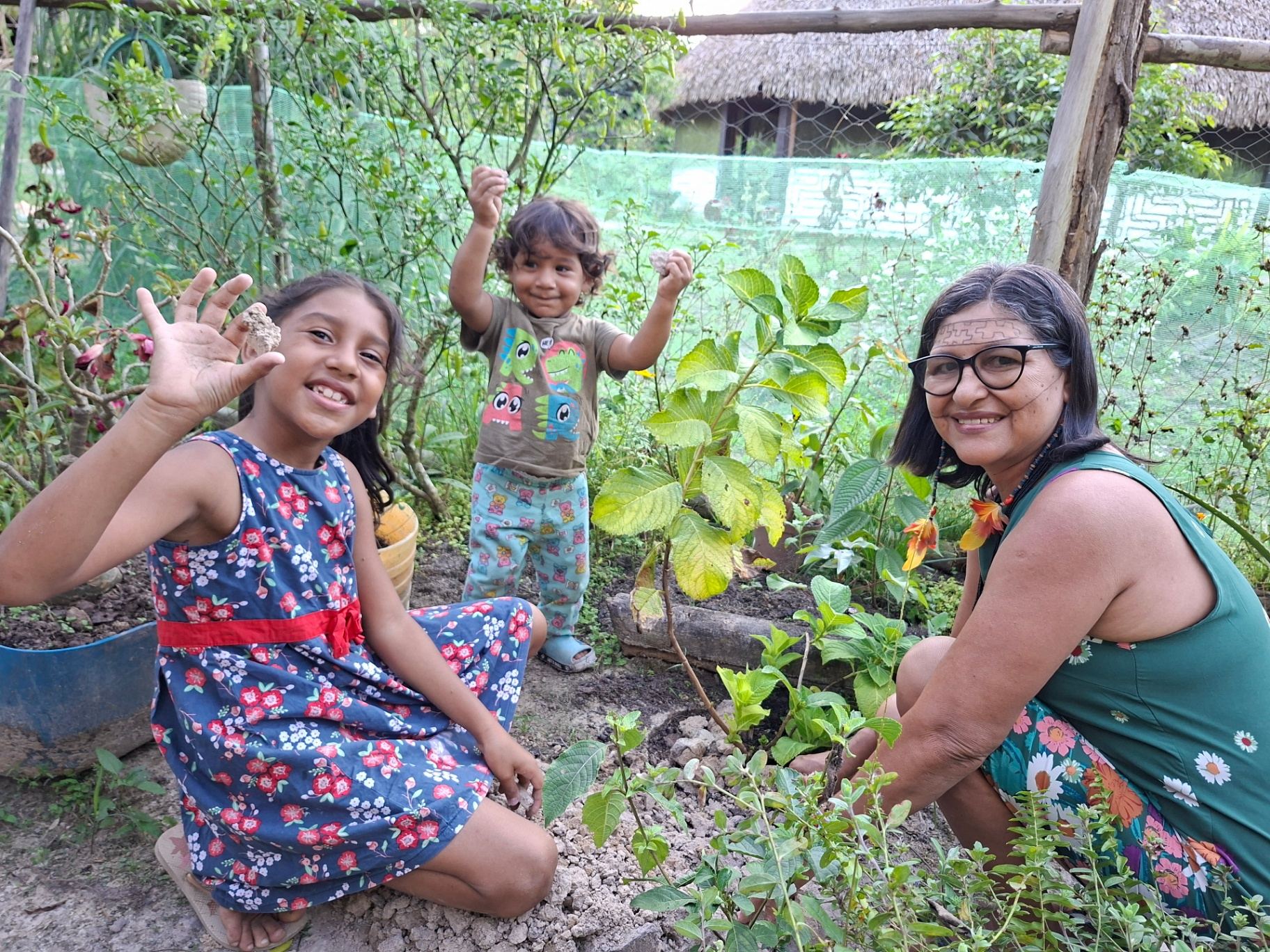 Maria Lucia Xipaia, elder and mother of Juma Xipaia, tending to plants in the Ipá-Supá medicinal herb garden alongside two smiling children in Kaarimã Village, Xipaya Indigenous Territory, Brazil.